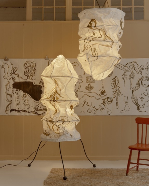 Two sculptural lampshades with artistic illustrations, standing and hanging in a room with art-covered walls and a red chair.