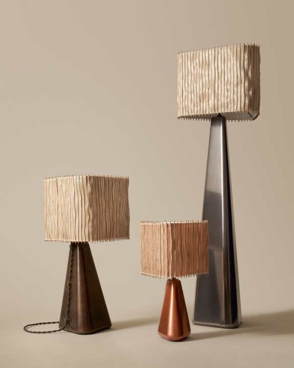 Three modern table lamps with pleated beige shades; bases in metallic finishes of bronze, copper, and silver on a neutral background.