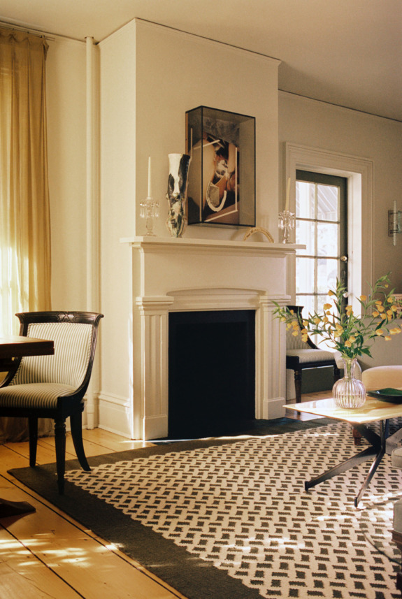 Elegant living room with a fireplace, patterned rug, wooden chairs, and a glass table, illuminated by natural light.