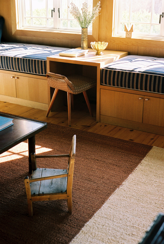 Cozy sunlit room with a wooden desk, chair, striped bench seating, vase with flowers, and a small wooden chair on a rug.
