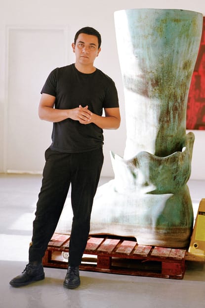 Person standing next to large sculpture in a gallery space with a pallet jack and abstract art on the wall in the background.