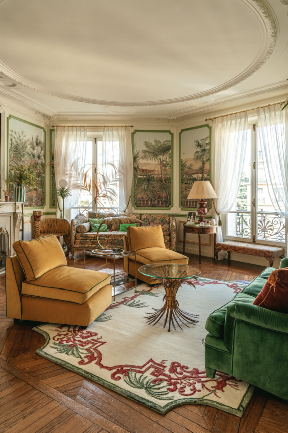 Elegant living room with vintage decor, featuring yellow chairs, green sofa, ornate rug, and large windows with light curtains.