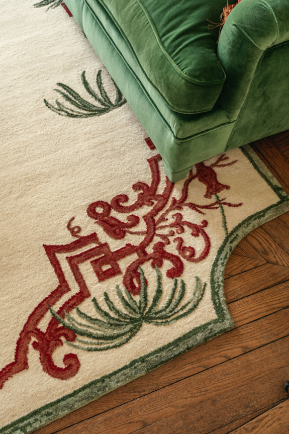 Vintage rug with red and green design on wooden floor next to a green velvet sofa.