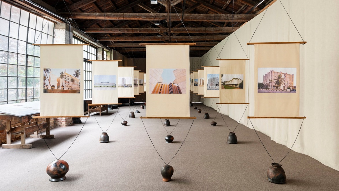 Art exhibition with hanging scrolls displaying architectural photographs in a well-lit room with large windows and wooden ceiling.