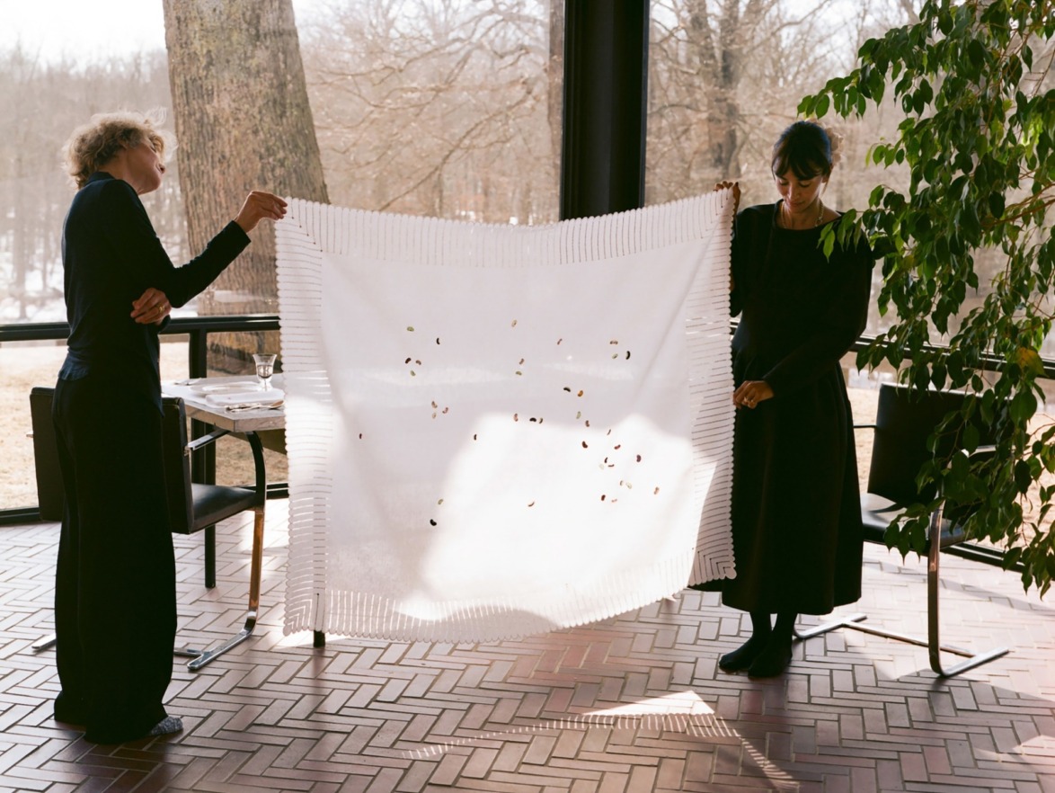 Two people holding a large embroidered fabric showcasing intricate designs, set in a room with large windows overlooking trees.