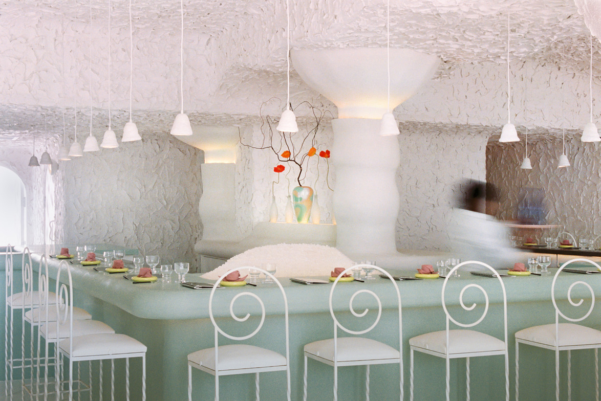 Modern restaurant interior with white textured walls, sleek bar, elegant chairs, and soft lighting.