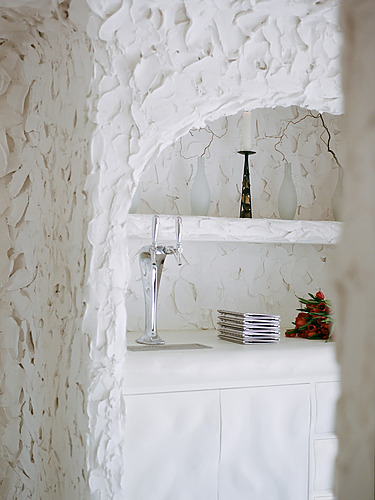 Elegant white plaster interior with niches, wine glasses, a candle, stacked coasters, and a bouquet of orange flowers.