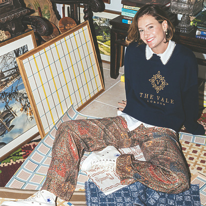 Person sitting on floor surrounded by patterned textiles, framed art, and books, wearing a navy sweater and colorful pants.