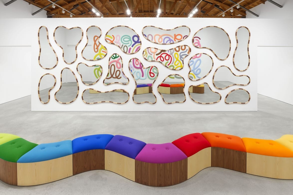 Colorful wavy bench in gallery with abstract wall art featuring wooden cutouts and vibrant swirling lines in the background.