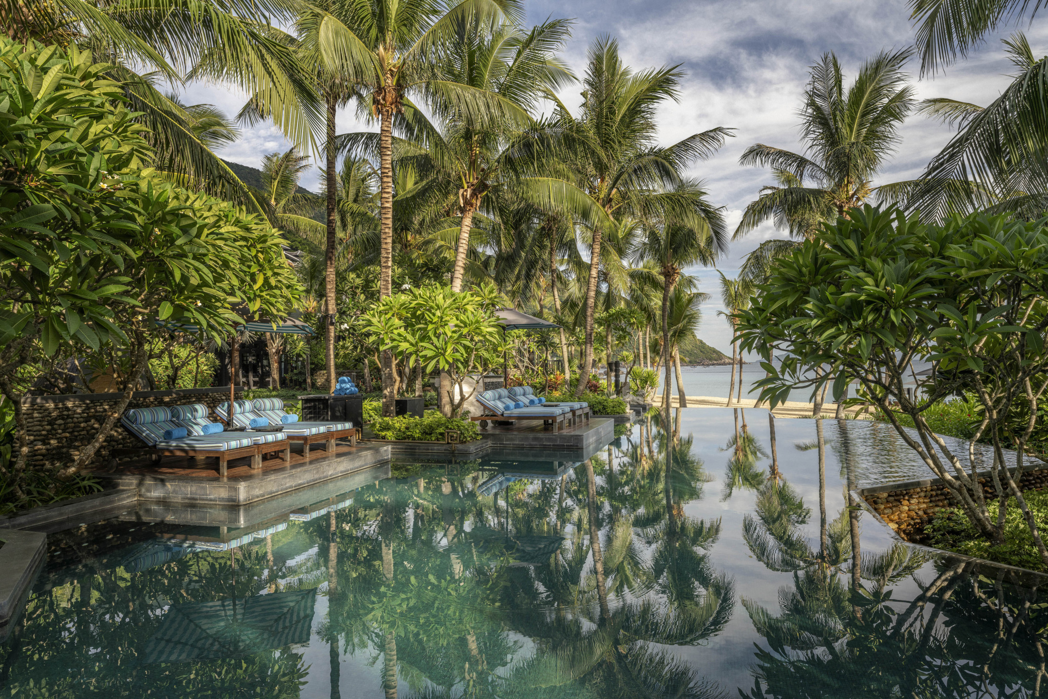 Luxurious resort pool surrounded by palm trees and lush greenery with a tranquil ocean view in the background.