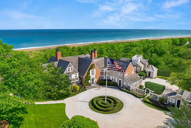 Aerial view of a large seaside mansion with an American flag, surrounded by lush greenery and overlooking a beach.