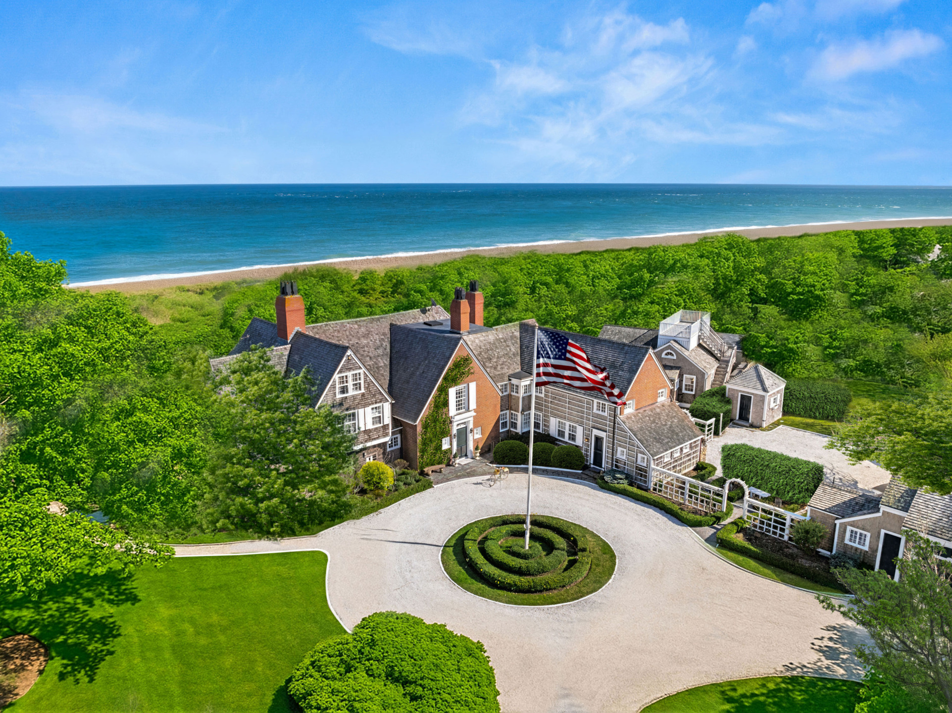 Aerial view of a large seaside mansion with an American flag, surrounded by lush greenery and overlooking a beach.