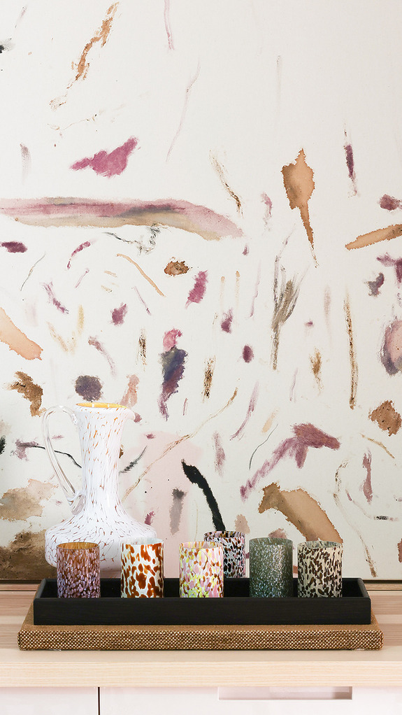 Abstractly painted wall, white ceramic pitcher, and a tray with six patterned candle holders on a wooden surface.