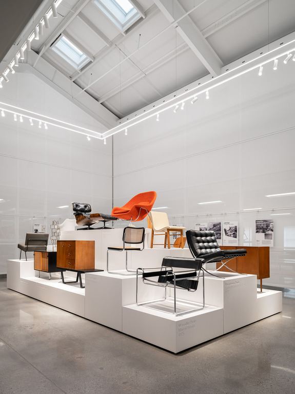 Exhibition display of modern chairs with various designs on white platforms in a well-lit room with high ceilings.