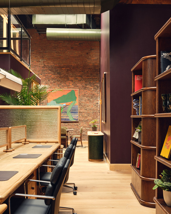 Modern coworking space with wood desks, black chairs, bookshelf, and colorful abstract art on exposed brick wall.