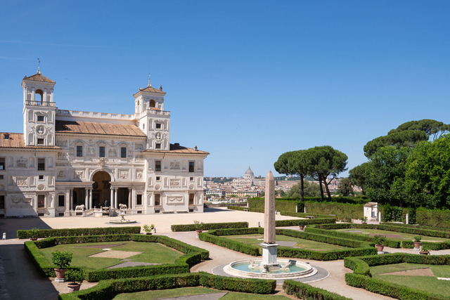 Villa with ornate facade and manicured gardens under a clear blue sky, surrounded by trees and distant cityscape in background.