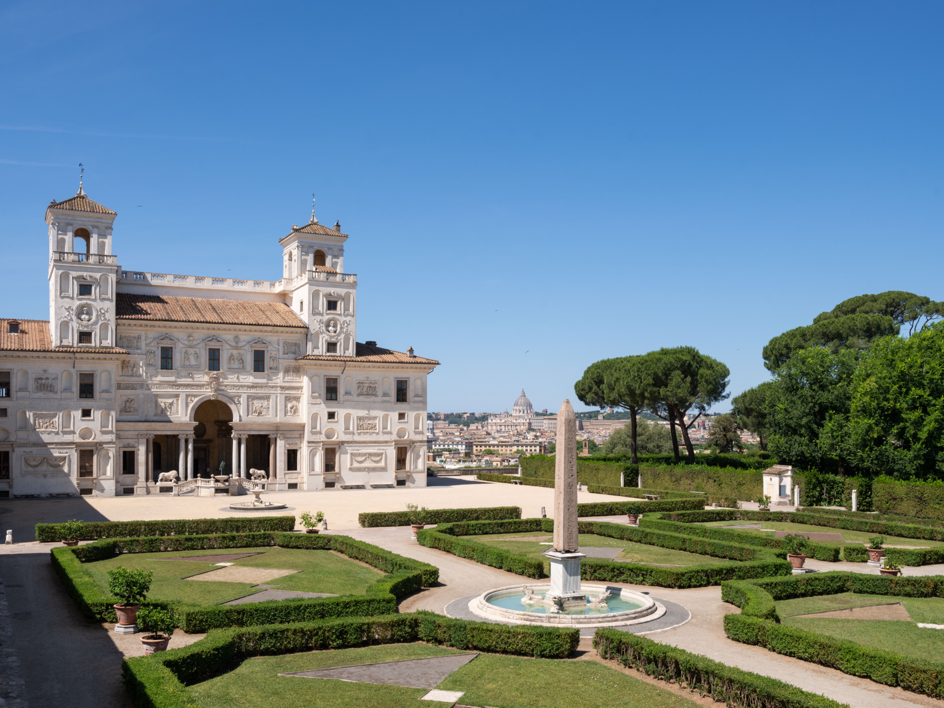 Villa with ornate facade and manicured gardens under a clear blue sky, surrounded by trees and distant cityscape in background.