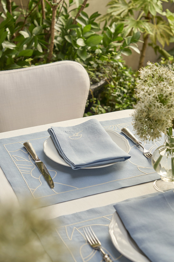 Elegant outdoor dining table setting with blue napkins, white plates, silver cutlery, and green plants in the background.