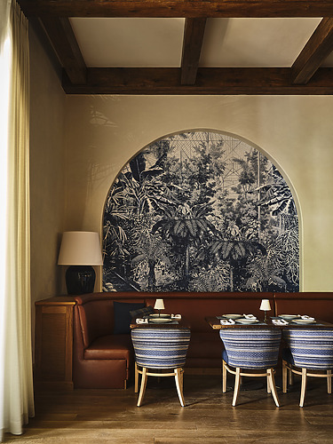 Cozy restaurant corner with two blue patterned chairs, a wooden table, and tropical themed wall mural under warm lighting.