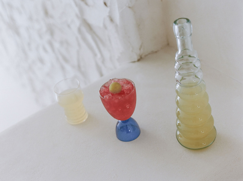 Three colorful drinks on a white surface: a tall drink, a red cocktail with ice and lemon slice, and a short drink in a glass