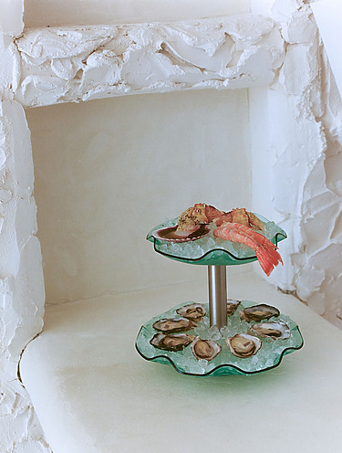Seafood platter with oysters, shrimp, and crab on a tiered glass stand with ice, set against a textured white background.