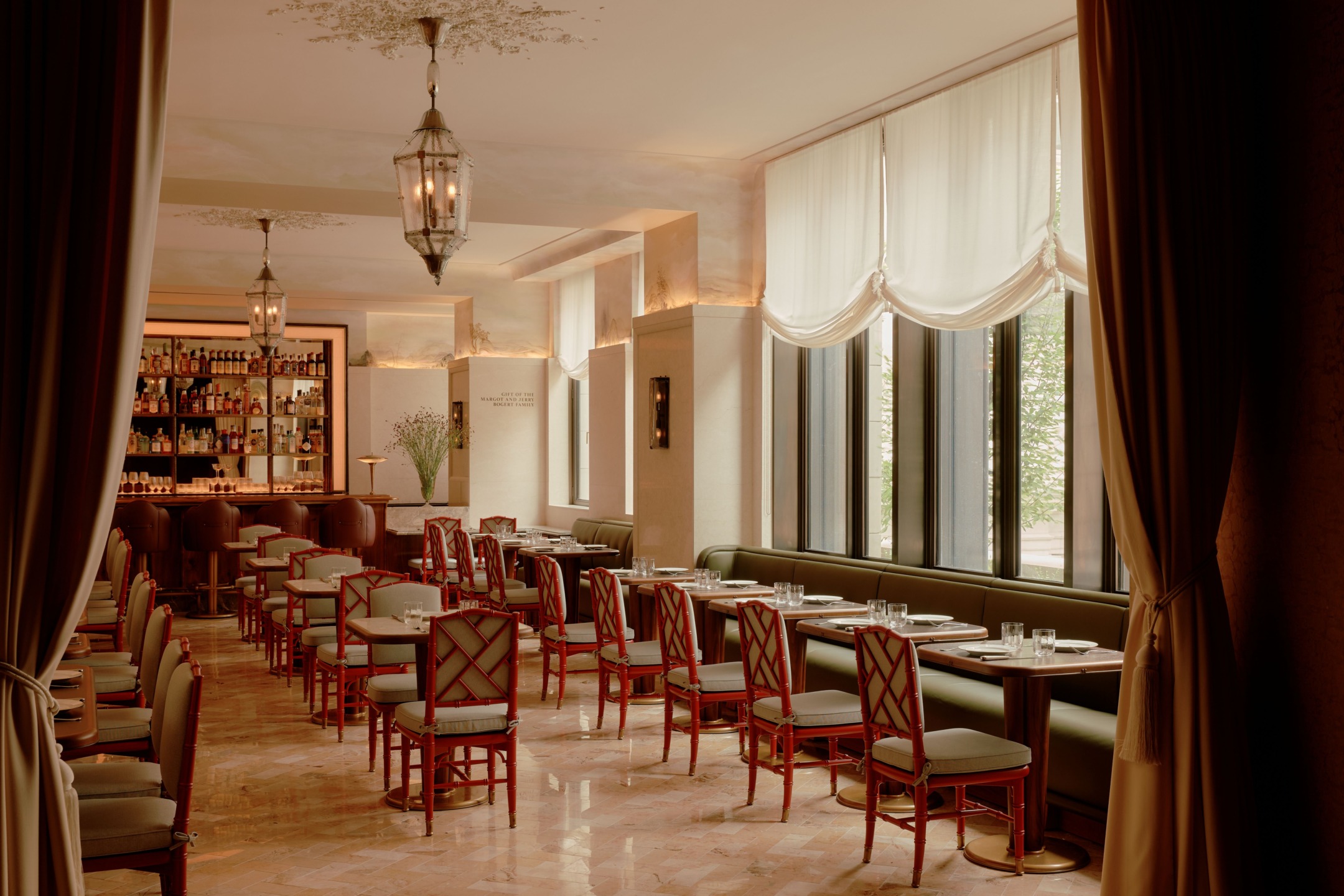 Elegant restaurant interior with neatly arranged dining tables, wooden chairs, a stocked bar, draped curtains, and soft lighting.