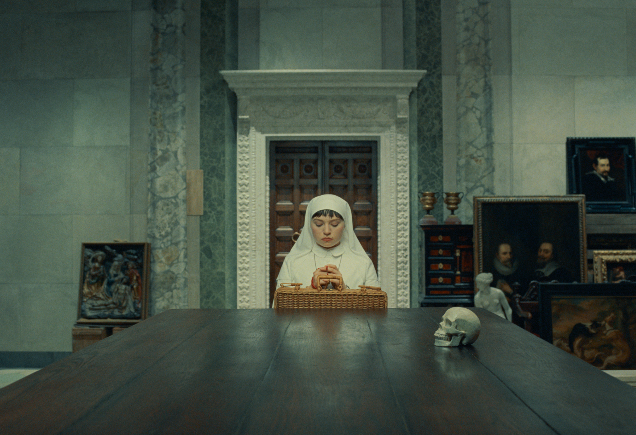 Nurse in white habit praying at a long table with a skull in an art-filled room.