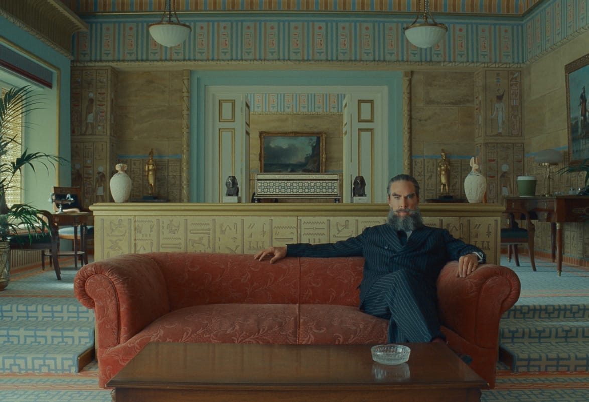 Man in a suit sitting on a red sofa in a luxurious room with ornate decor and a large decorative wall in the background.