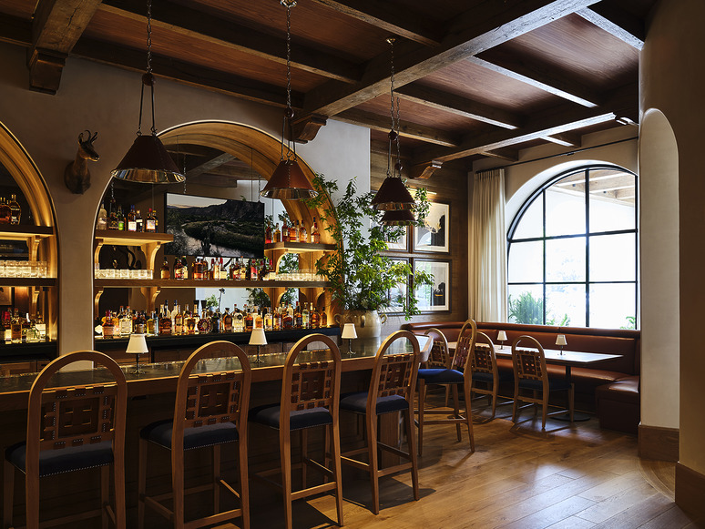 Cozy bar interior with wooden stools, arched shelves displaying bottles, pendant lights, and large arched window.