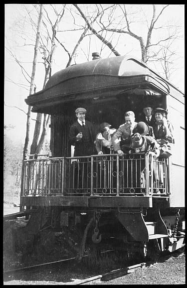 People gathered on the platform of a vintage train car in a wooded area, wearing early 20th-century clothing.