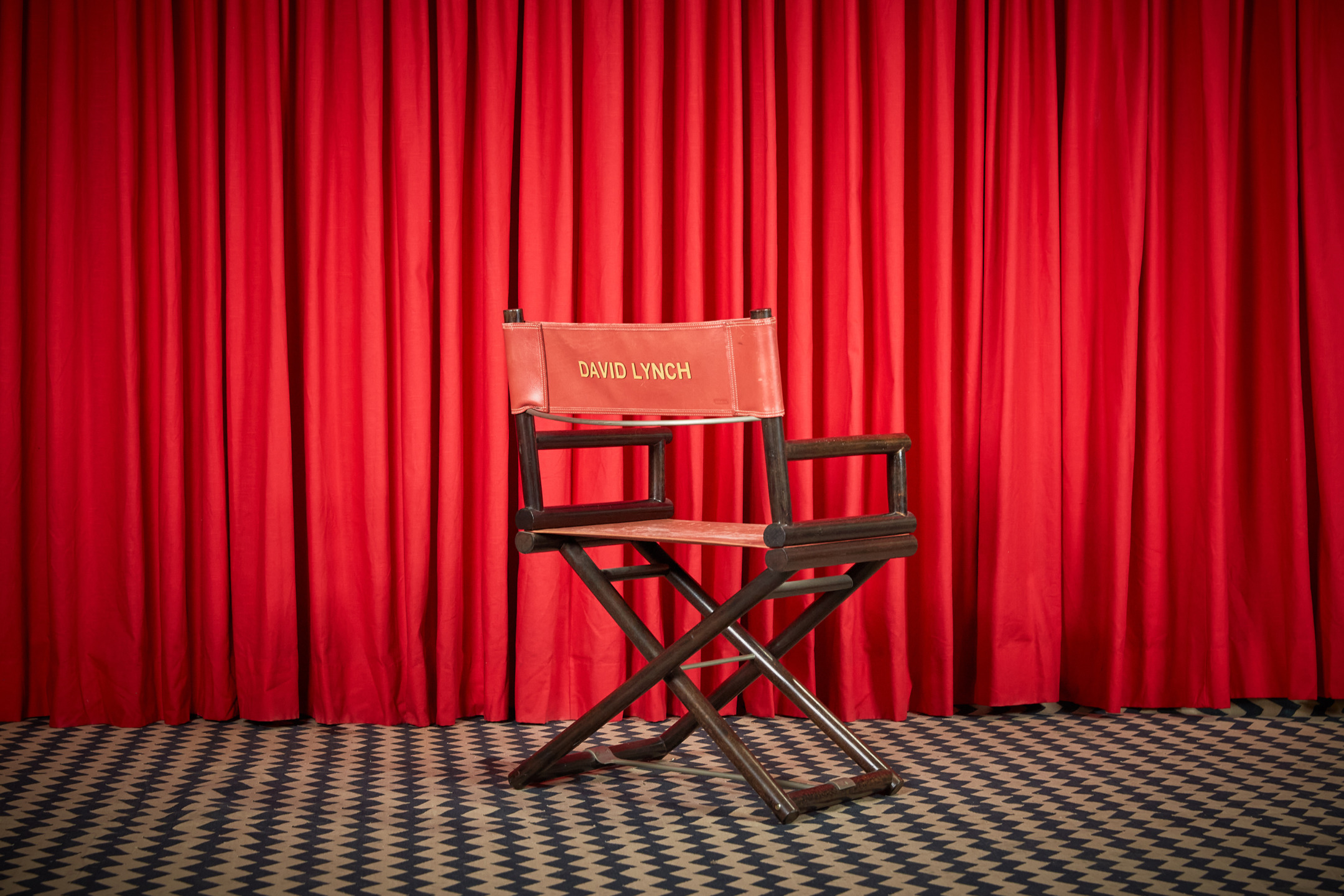 Director's chair with red fabric and wooden frame in front of matching red theater curtain and checkered floor.