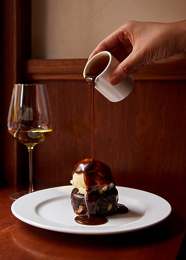 Pouring chocolate sauce over ice cream-topped brownie on white plate, with a glass of white wine in the background.