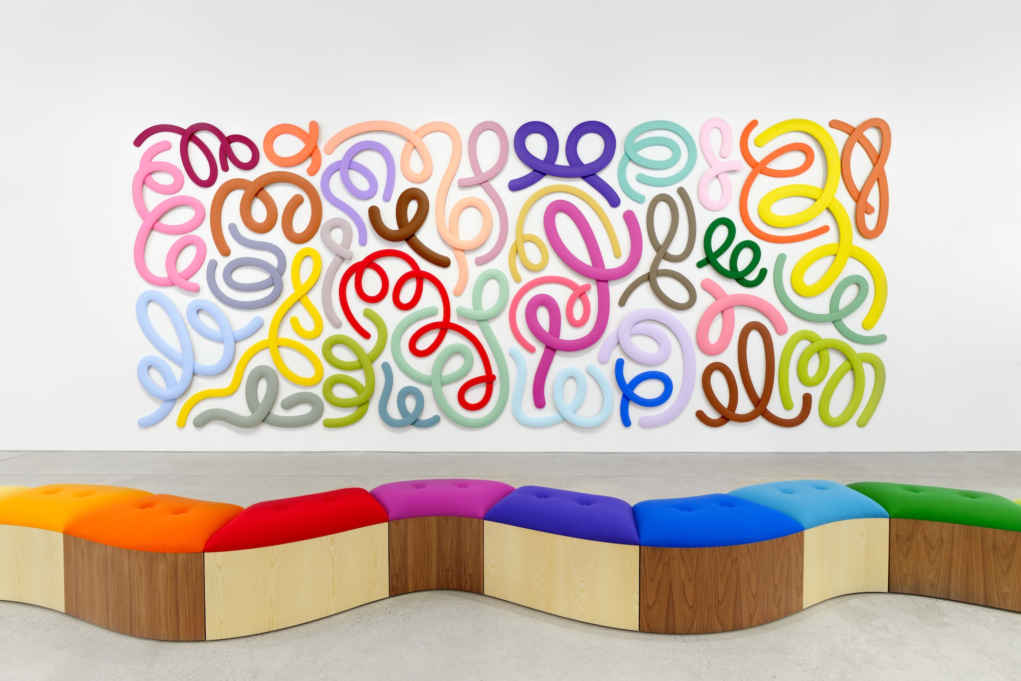 Colorful abstract wall art with swirling patterns above a wavy, multicolored bench in a modern gallery setting.