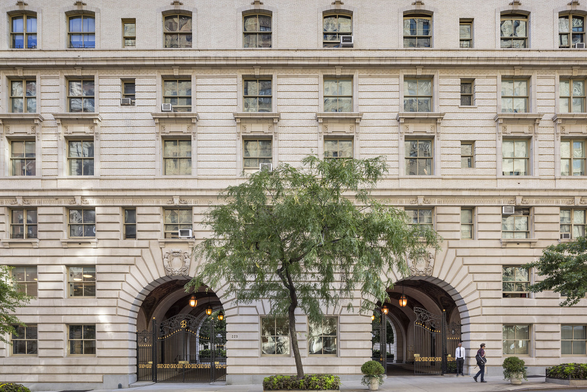 Historic brick building facade with arched entrance and windows, tree and pedestrians in foreground during daytime.
