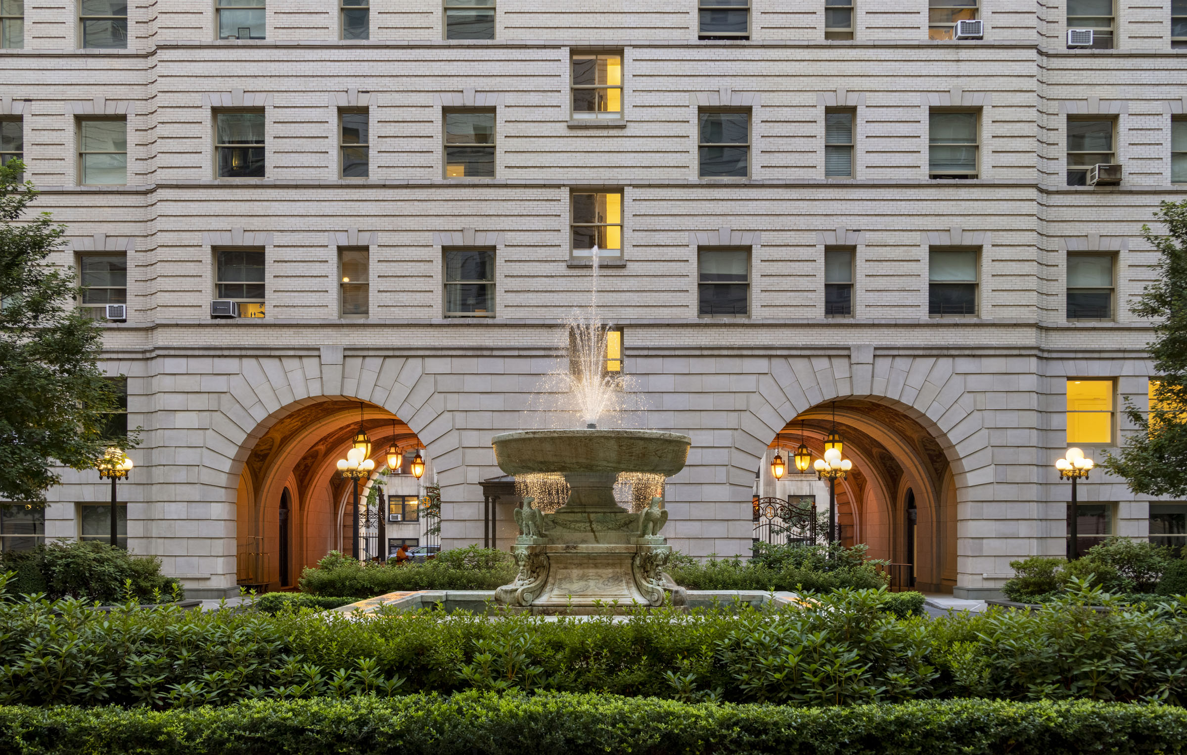 Fountain in front of a historic building with arched windows and lights, surrounded by green bushes and trees.