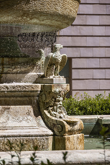 Stone sculpture of an eagle on a decorative fountain edge with water flowing, surrounded by green bushes and a brick wall.