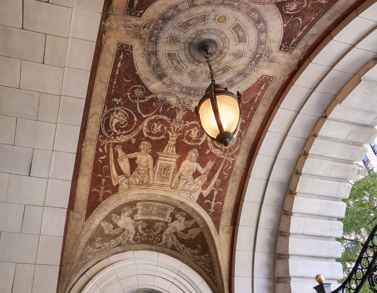 Ornate grand archway with detailed artwork, depicting figures holding shields and a central light fixture, surrounded by decorative tiles.