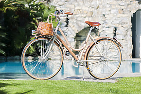 Vintage-style bicycle with brown leather seat and basket parked on green grass beside a pool and stone wall in sunny weather.