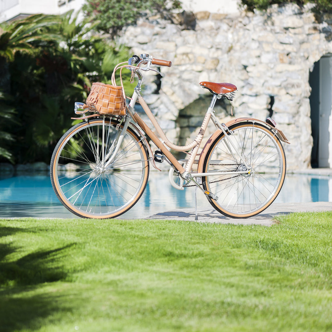 Vintage-style bicycle with brown leather seat and basket parked on green grass beside a pool and stone wall in sunny weather.