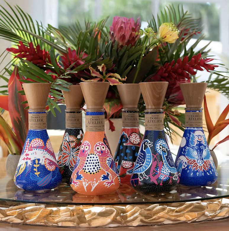 Colorful decorative tequila bottles with intricate designs on a table, surrounded by vibrant tropical flowers.