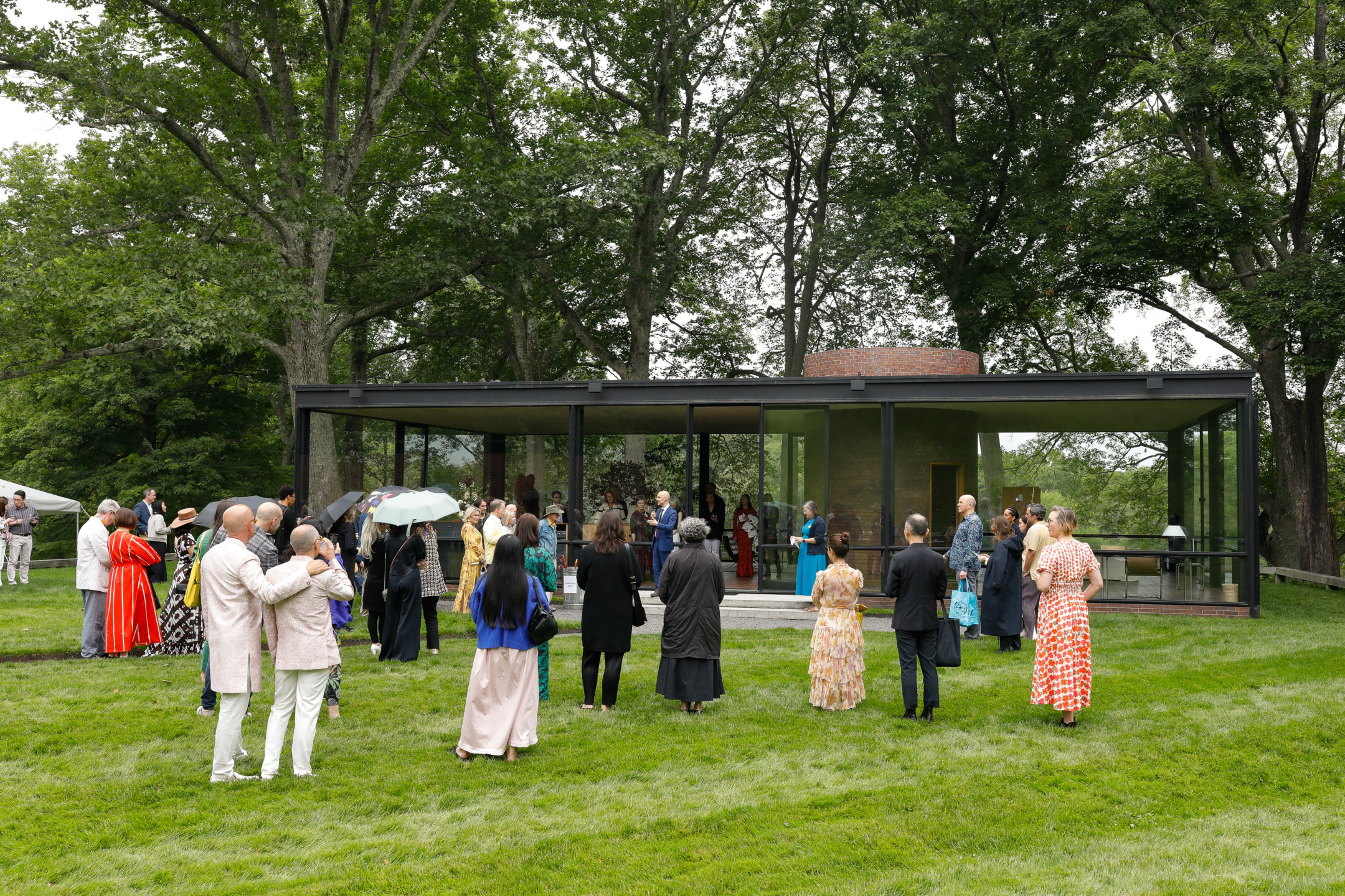 A group of people gathered outside a modern glass building surrounded by trees and grass on a cloudy day.
