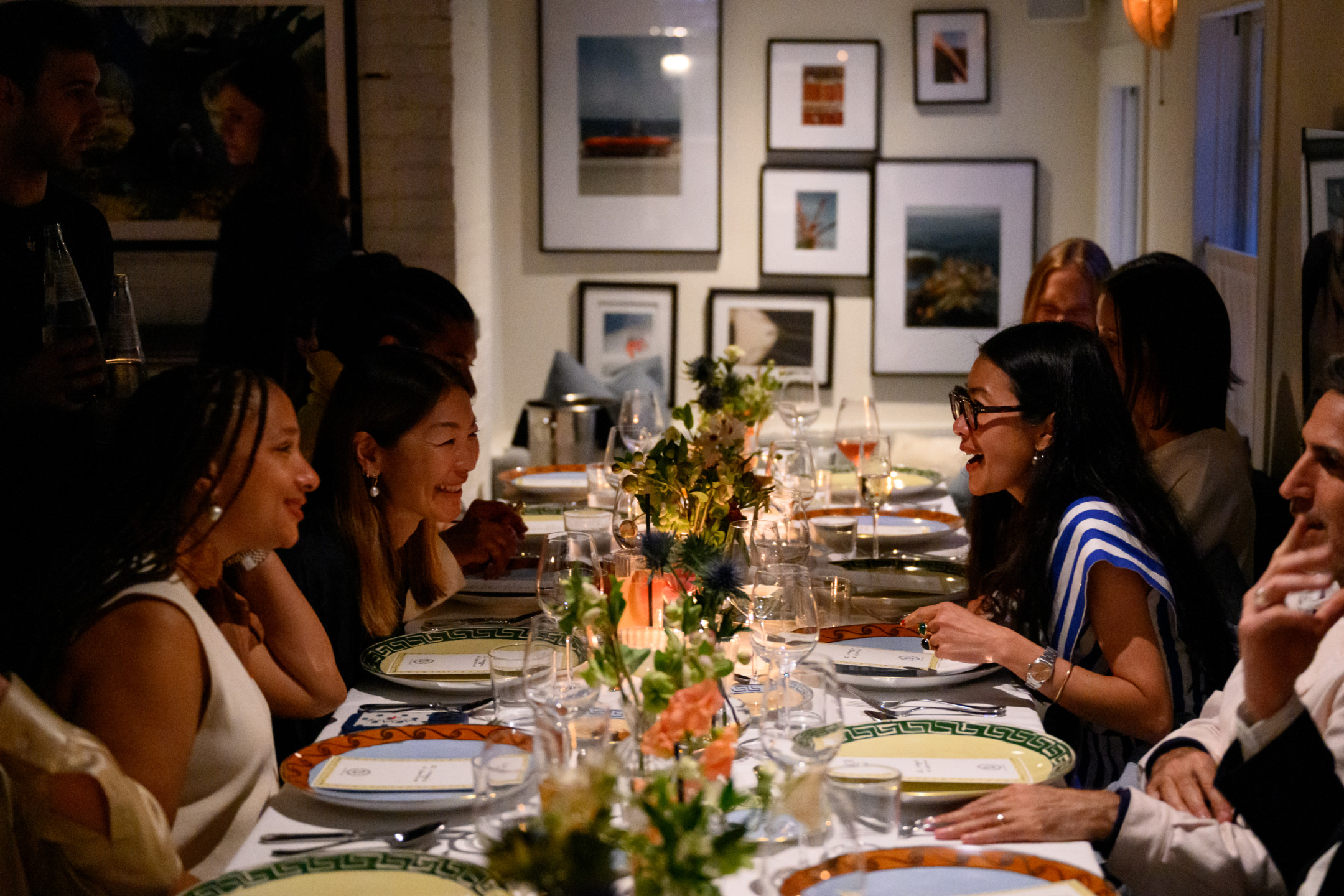 People enjoying a lively dinner party with elegant table settings and artwork on the walls in a cozy, dimly lit room.