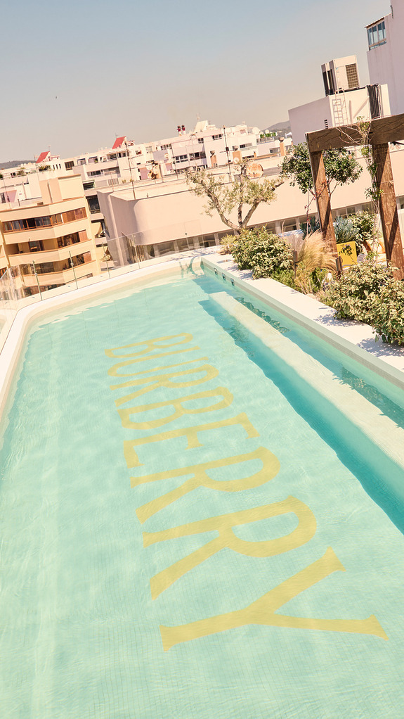 Rooftop pool with skyline view, featuring large BURBERRY logo on clear blue water.