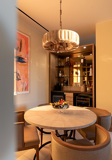 Dining area with round table, chairs, large chandelier, and lit cabinet with assorted decorations and bottles