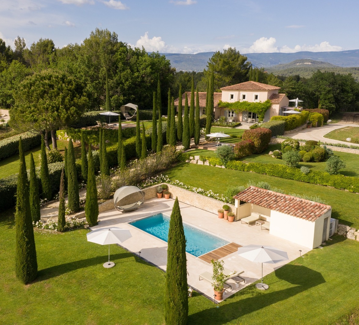 Aerial view of a villa with a pool, surrounded by lush greenery, trees, and mountains in the background.