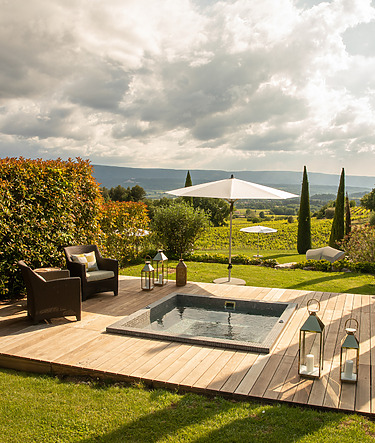 Wooden deck with a small hot tub, lounge chairs, umbrella, and scenic view of lush greenery and cloudy sky.