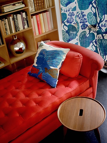 Red chaise lounge with a colorful pillow, next to a wooden side table, bookshelves, and a floral-patterned curtain in the background.