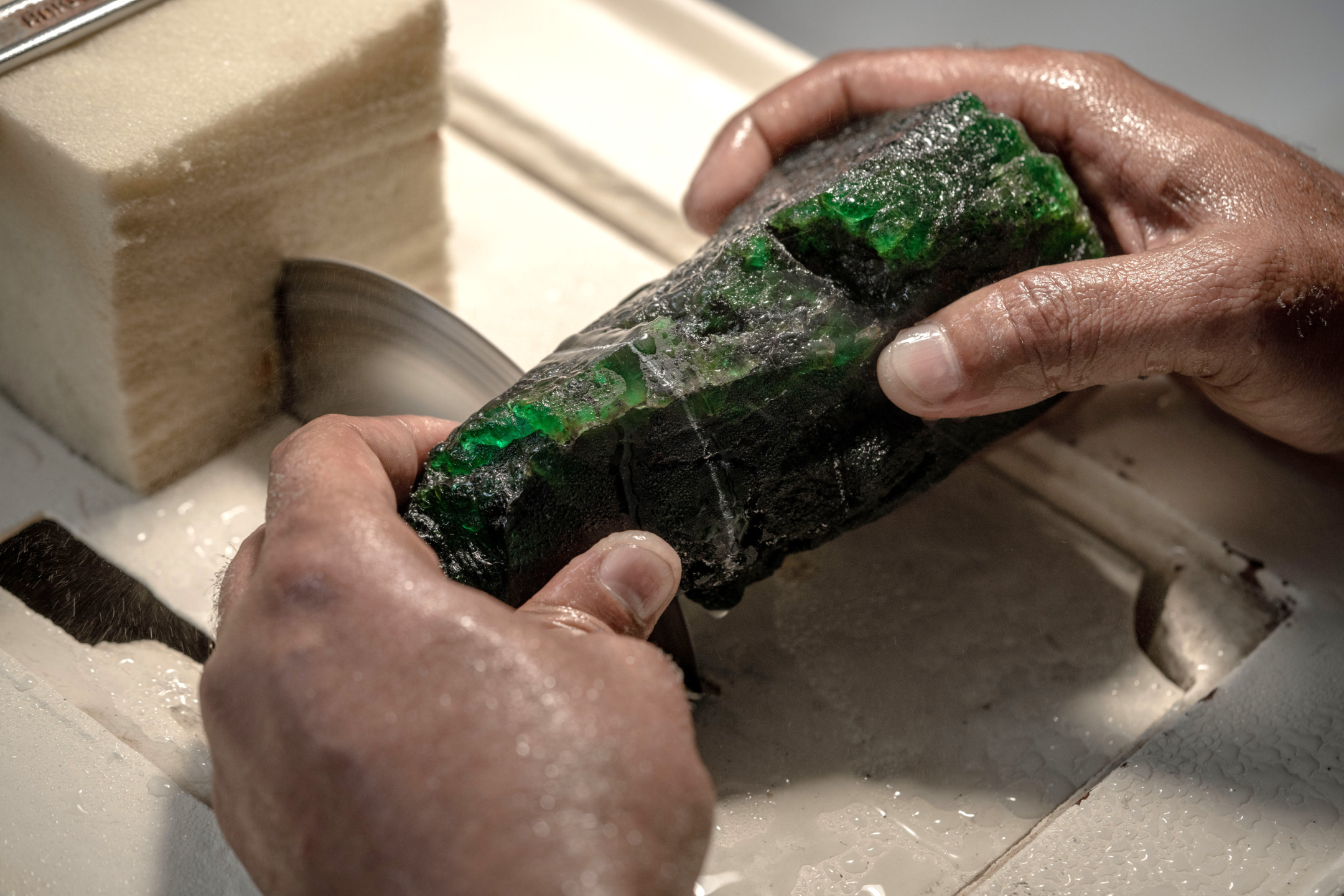 Person cutting a large, rough emerald stone with a circular saw, focusing on precision and detail.