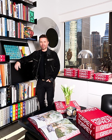 Person in a room with bookshelves, books titled "TORREY," a clock, and a view of city buildings through the window.