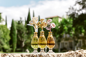 Three gold glass vases with decorative flowers on a stone ledge, green trees and blurred house in the background.